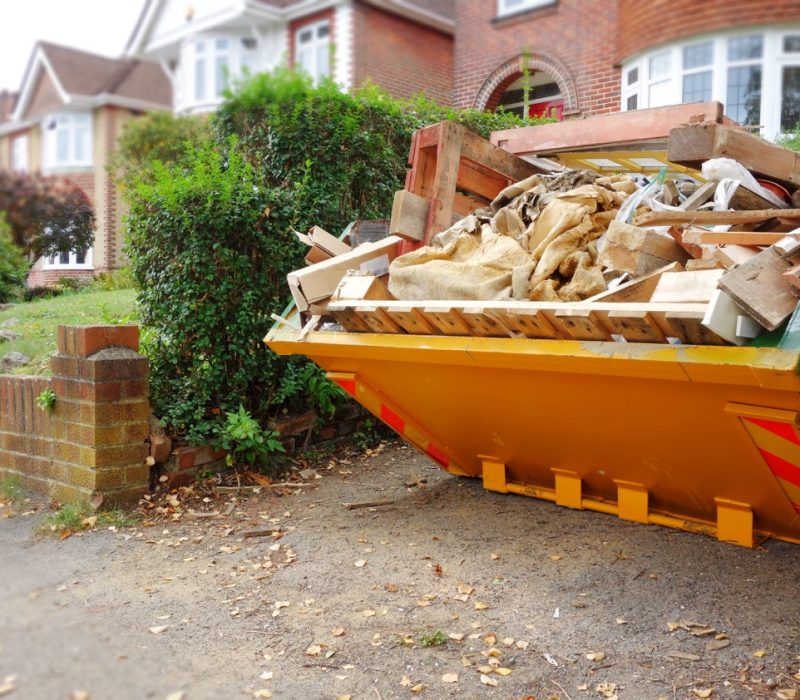 Yellow,Rubbish,Skip,On,Driveway.,Selective,Focus,On,Full,Skip Yellow rubbish skip on driveway. Selective focus on full skip with space to add text in front of bin, footpath & blurry background of green bush fence, houses, brick wall. Renovate, moving concept.
